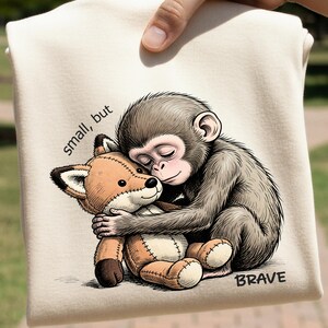 Op de afbeelding: Een beige t-shirt met een cartoon illustratie van een aap die een vossenknuffel omhelst. De tekst "small, but BRAVE" staat op het shirt. De aap is grijs en de vos is bruin en beige.