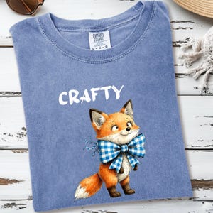 May include: A blue Comfort Colors t-shirt with the word "CRAFTY" in white. The shirt features a cartoon fox wearing a blue and white checkered bow tie. The fox is orange, white, and brown.