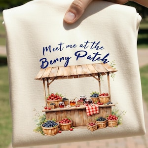 May include: Cream-colored sweatshirt featuring a watercolor illustration of a berry stand. The stand is filled with baskets of berries, jars of jam, and the text "Meet me at the Berry Patch" in blue script.