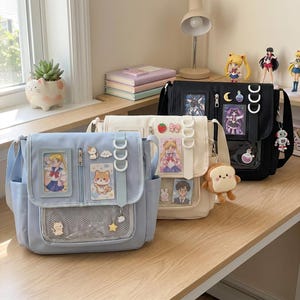 May include: Three colorful messenger bags in blue, cream, and black. Each bag features a flap closure, multiple pockets, and decorative charms. The bags have a mesh window and are adorned with anime-style character cards and cute accessories.