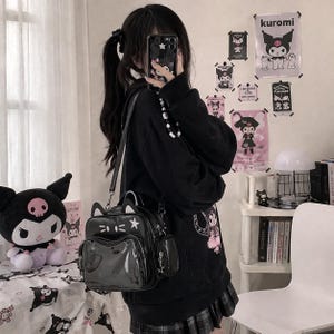 Puede incluir: Mochila negra con orejas de gato, una estrella y un bolsillo frontal transparente. La bolsa tiene un pequeño bolsillo lateral y se usa con una sudadera con capucha negra y una falda a cuadros. Hay carteles de un personaje de dibujos animados en la pared.