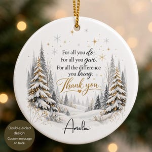 May include: A round white ceramic ornament featuring a winter forest scene with snow-covered pine trees, gold star accents, and the text: For all you do. For all you give. For all the difference you bring. Thank you. The name Amelia is printed below.