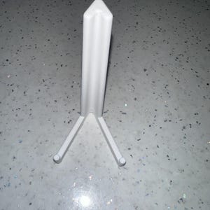May include: A white, triangular stand with three legs. The stand is made of a solid material and is designed to hold an item. The stand is on a speckled surface.