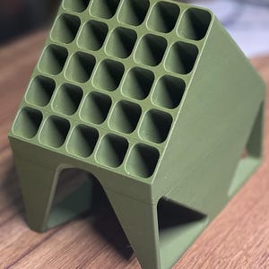 May include: A green, 3D-printed organizer with 25 square compartments. The angled design features a grid of evenly spaced holes, supported by a base with triangular cutouts. The object is made of a solid, matte green material.