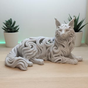 Op de afbeelding: Een lichtgrijs, driedimensionaal kattenbeeldje met een ingewikkeld, opengewerkt filigraanontwerp dat vloeiende, wervelende vachtpatronen nabootst. De kat is afgebeeld in een rustende, liggende positie met de kop rechtop en alert.