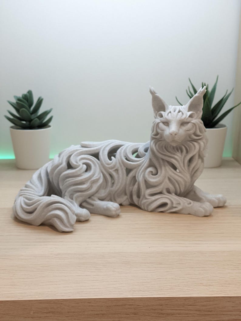 Op de afbeelding: Een lichtgrijs, driedimensionaal kattenbeeldje met een ingewikkeld, opengewerkt filigraanontwerp dat vloeiende, wervelende vachtpatronen nabootst. De kat is afgebeeld in een rustende, liggende positie met de kop rechtop en alert.