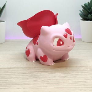 May include: A light pink creature figurine with a large red rose-shaped bulb on its back. The creature features red heart-shaped spots on its forehead and sides, with white and red eyes. The surface has a matte texture and a smooth finish.