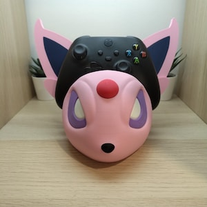 Può includere: Un supporto per controller a forma di creatura rosa e viola che sostiene un controller di gioco wireless nero. Il supporto presenta grandi orecchie a punta con accenti blu scuro, una gemma frontale rossa, occhi bianchi e un piccolo naso nero.