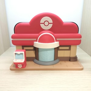 Könnte beinhalten: Ein 3D-gedrucktes Modell eines Pokémon Centers. Das Gebäude hat ein rotes Dach mit einem weißen und roten Pokéball-Emblem, beige Wände und eine hellblaue Tür. Auf der linken Seite befindet sich eine kleine rote Maschine mit einem Bildschirm.