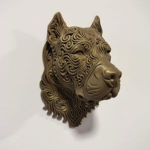 Könnte beinhalten: Eine bronzefarbene Wandskulptur eines Hundekopfes mit komplexen, erhabenen Wirbelmustern auf der gesamten Oberfläche. Die Hunde-Figur besitzt spitze Ohren, eine ausgeprägte Schnauze und eine texturierte Mähne, die sich vom Hals abwärts erstreckt.