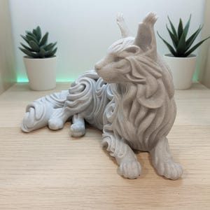 Op de afbeelding: Een lichtgrijs, mat afgewerkt beeldje van een liggende kat met gestileerde, vloeiende vachtpatronen. Het beeldhouwwerk heeft puntige oren met pluimpjes, een slanke katachtige houding en een getextureerd oppervlak dat op steen lijkt.
