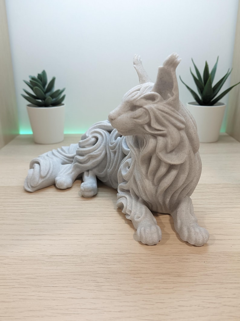 Op de afbeelding: Een lichtgrijs, mat afgewerkt beeldje van een liggende kat met gestileerde, vloeiende vachtpatronen. Het beeldhouwwerk heeft puntige oren met pluimpjes, een slanke katachtige houding en een getextureerd oppervlak dat op steen lijkt.