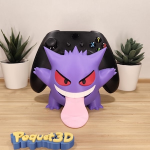 May include: A purple Gengar character controller stand holding a black game controller. The character has red eyes, a wide white-toothed grin, and a long pink tongue extending downward. A blue and yellow sign with the text Poquet3D is in front.