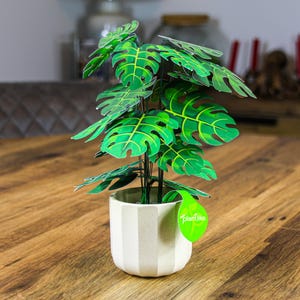May include: A paper plant in a white and beige striped pot. The plant has large green leaves with yellow veins and brown accents. A green tag with the text "Plantlike" is attached to the plant.
