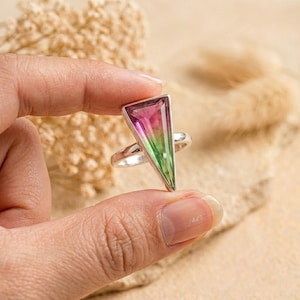 May include: A silver ring featuring a large, triangular-shaped watermelon tourmaline gemstone. The stone displays a distinct color gradient, transitioning from a vibrant pink at the top to a bright green at the base, set within a polished metal frame.