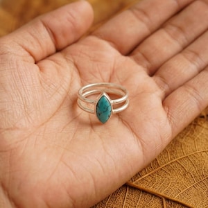 Op de afbeelding: Een zilveren ring met een dubbel bandontwerp, met een turquoise marquise-geslepen steen. De ring wordt op een hand getoond, waarbij de details en het kleurcontrast van de sieraden worden getoond. Modieuze sieraden.