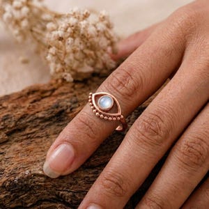 Peut inclure: Une bague en or rose présentant un motif en forme d'œil avec un cabochon de pierre de lune rond au centre. L'anneau en métal est orné de petits détails perlés le long du bord extérieur de la forme d'œil, créant une finition texturée sur le bijou.