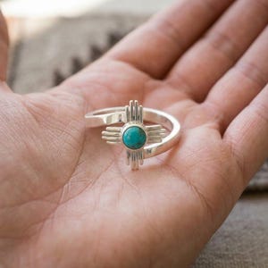 Op de afbeelding: Een zilveren ring met een centrale ronde turkoois steen gezet in een kruisvormig ontwerp met vier uitstralende armen, elk bestaande uit drie parallelle verticale staven, bevestigd aan een gladde, gepolijste zilveren ringband.