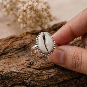 May include: A silver ring with a white and black cowrie shell. The shell is oval and set within a beaded silver border. The ring is shown on a wooden surface.