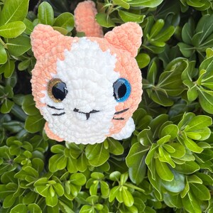 May include: A handmade crochet cat toy with peach and white yarn. The cat has one gold and one blue eye, with black stitched details for the nose and whiskers. The cat is set against a background of green leaves.