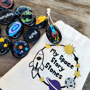 May include: A white drawstring bag with the text "My Space Story Stones" printed on it. The bag is filled with black and white painted stones with space-themed designs, including planets, stars, and a rocket.