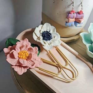 May include: Three gold paper clips with a pink flower and a white flower attached to them. The flowers are made of polymer clay and have a realistic appearance. The paper clips are on a light pink tray.
