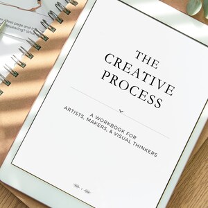 May include: A white tablet showing the title "THE CREATIVE PROCESS" in black lettering. Underneath, it states "A WORKBOOK FOR ARTISTS, MAKERS, & VISUAL THINKERS." The tablet is on a brown surface with a spiral notebook and glasses.