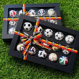 Könnte beinhalten: Zwei schwarze Präsentationsboxen gefüllt mit Miniaturfußbällen. Jede Box ist mit einem Band mit Fußbällen und den Farben Rot, Schwarz und Gelb verziert. Die Boxen stehen auf einer grünen, grasbewachsenen Oberfläche.