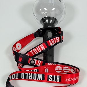 Puede incluir: Un bastón luminoso BTS negro con una parte superior esférica transparente que muestra el logo de BTS. Se adjunta un cordón rojo y negro, impreso con el texto BTS WORLD TOUR, ARMY y los nombres de miembros como J-HOPE, JUNG KOOK y RM en blanco.