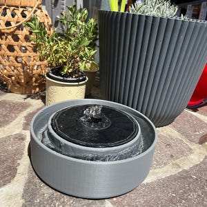 Pode incluir: Uma fonte de &aacute;gua circular cinzenta com energia solar, um painel solar preto no centro e um pequeno bico de &aacute;gua. A fonte est&aacute; cheia de &aacute;gua e assenta numa superf&iacute;cie de pedra, em frente a um vaso cinzento canelado e um lanterna tecida.