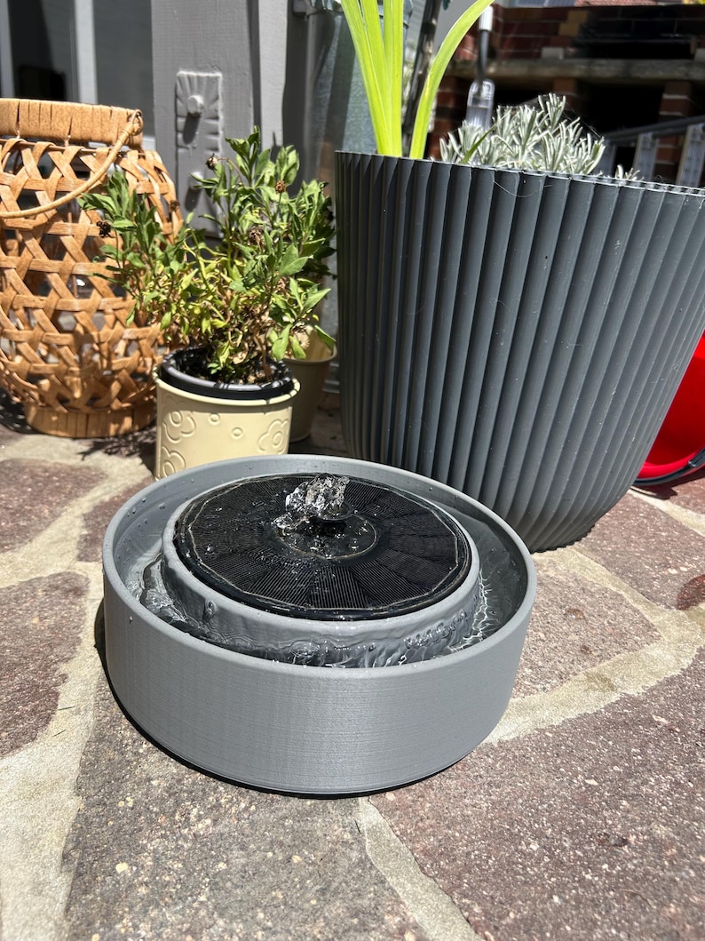 Pode incluir: Uma fonte de &aacute;gua circular cinzenta com energia solar, um painel solar preto no centro e um pequeno bico de &aacute;gua. A fonte est&aacute; cheia de &aacute;gua e assenta numa superf&iacute;cie de pedra, em frente a um vaso cinzento canelado e um lanterna tecida.