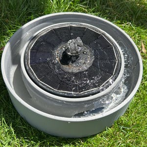 Pode incluir: Uma bacia circular cinzenta contendo uma fonte de &aacute;gua flutuante alimentada por energia solar. A fonte possui um painel solar circular preto com um bocal central que pulveriza um pequeno jato de &aacute;gua para cima.