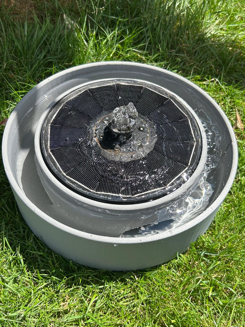 Pode incluir: Uma bacia circular cinzenta contendo uma fonte de &aacute;gua flutuante alimentada por energia solar. A fonte possui um painel solar circular preto com um bocal central que pulveriza um pequeno jato de &aacute;gua para cima.