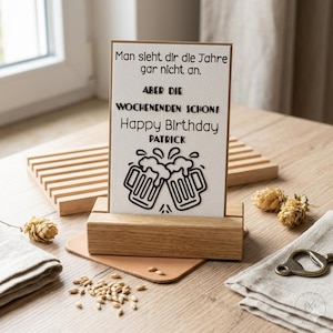 May include: A birthday card with the text "Man sieht dir die Jahre gar nicht an. ABER DIE WOCHENENDEN SCHON! Happy Birthday PATRICK" and two beer mugs. The card is in a wooden holder on a wooden table.