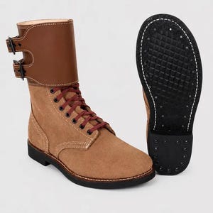 Puede incluir: Botas de combate de ante color canela con detalles de cuero marrón y cordones burdeos. Las botas tienen un diseño de caña alta, dos cierres de hebilla y una suela negra. Una bota se muestra con la suela hacia arriba.