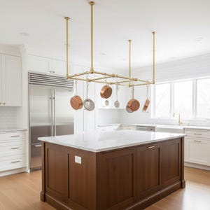 May include: A rectangular brass pot rack suspended from the ceiling above a kitchen island. The rack features a grid frame with multiple hooks holding various copper and stainless steel cookware, including round pans and small kitchen utensils.