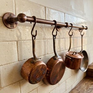 May include: Copper kitchen pot rack with hanging pots and pans. The rack is mounted on a tiled wall. The pots and pans are copper with brass handles. The rack is made of copper. The kitchenware is vintage.