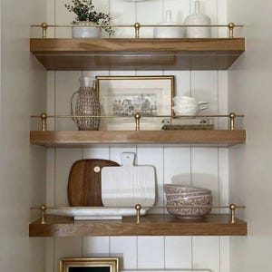 May include: Wooden shelves with brass accents display decorative items. A framed picture, cutting boards, vases, and a small plant are arranged on the shelves. A gold-toned wall sconce is mounted above the top shelf. The overall aesthetic is a blend of rustic and elegant.