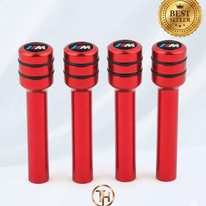 May include: Four red car door lock pins with the BMW M logo on top. The pins are cylindrical with black accent rings. A gold "Best Seller" sticker is in the upper right corner. The Trendy Hub logo is at the bottom.