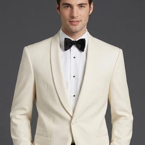 May include: An off-white dinner jacket featuring a shawl lapel and a single-button closure, worn over a crisp white dress shirt with a black bow tie. The jacket includes two side pockets and is tailored with long sleeves and structured shoulders.