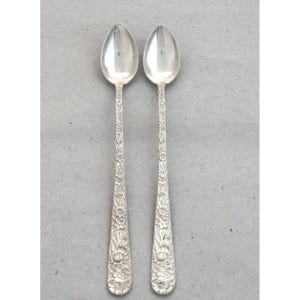May include: Two silver spoons with floral detailing. The spoons have a classic design with a rounded bowl and a long handle adorned with intricate flower patterns. The spoons are likely made of sterling silver.