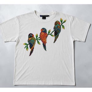 Può includere: Una t-shirt bianca a maniche corte con una stampa grafica di tre pappagalli colorati appollaiati su un ramo verde con foglie. I pappagalli sono raffigurati in tonalità arancione, blu e verde. L'etichetta del marchio I.B. DIFFUSION è visibile sul collo.