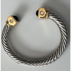 May include: A silver-toned, twisted cable cuff bracelet with gold-colored accents. The bracelet features a black stone and a small, clear crystal on each end. The ends are capped with gold-colored metal.
