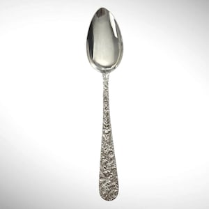 May include: A silver-toned metal spoon featuring a smooth, polished oval bowl and a handle intricately embossed with a dense, continuous pattern of small flowers, leaves, and stems covering the entire length of the handle surface.