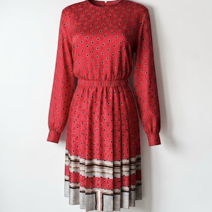May include: A red dress with long sleeves and a round neckline. The dress features a polka dot pattern and a pleated skirt with a geometric border in black, white, and tan. The waist is cinched with an elastic band.