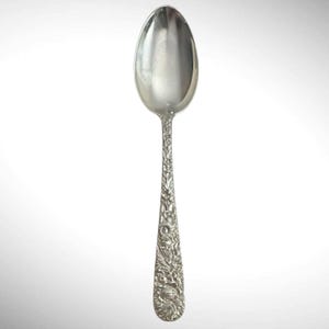 Op de afbeelding: Een zilverkleurige metalen lepel met een ingewikkeld, in reliëf aangebracht bloemen- en bladpatroon dat de gehele lengte van de steel bedekt. De lepelbak is glad, gepolijst en reflecterend, wat contrasteert met het gedetailleerde, textuurrijke ontwerp.