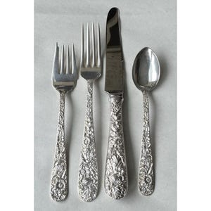 Pode incluir: Um conjunto de quatro talheres prateados: um garfo, uma faca, uma colher e um garfo de salada. Cada peça apresenta um padrão floral no cabo. A faca tem uma lâmina curva. Os talheres estão dispostos sobre uma superfície cinza claro.