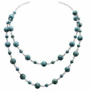 Puede incluir: Un collar de doble hebra con cuentas esféricas de color turquesa de varios tamaños. Las cuentas están ensartadas en una cadena de color plateado, creando un efecto de capas. El collar se muestra sobre un fondo blanco.