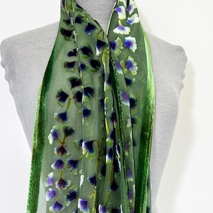 May include: A sheer green scarf with a floral pattern of purple and white flowers. The scarf has a green velvet border and is draped over a mannequin. The scarf is a fashion accessory.