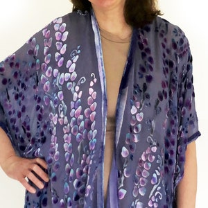 Op de afbeelding: Een doorschijnende, open kimono in tinten paars en blauw, versierd met een bloemmotief. De kimono heeft korte mouwen en is afgezet met een donkerder paars fluweelachtig materiaal. Het wordt gedragen over een neutraal gekleurde jurk.